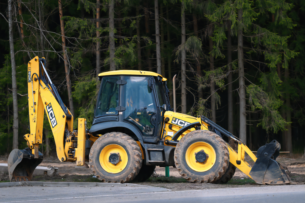 Koparka JCB 4CX ECO stojąca przy drodze, otoczona drzewami.