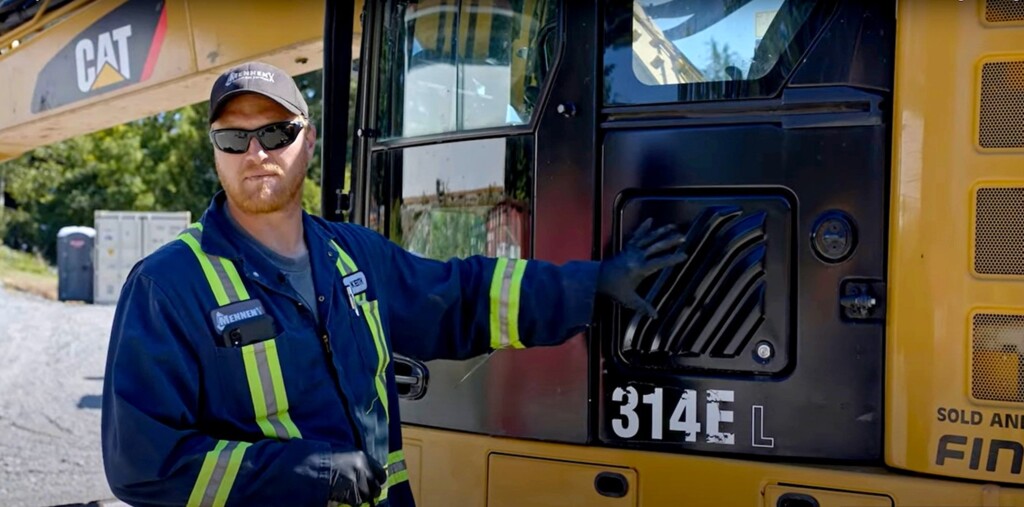 Mechanik w odzieży ochronnej stoi przy koparce CAT 314E L na placu budowy.