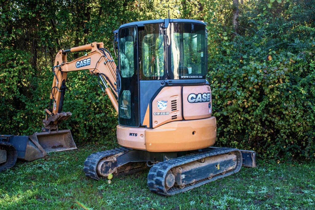Obrázek ukazuje malý pásový bagr značky CASE, model CX36B, zaparkovaný na trávě vedle hustého křoví.