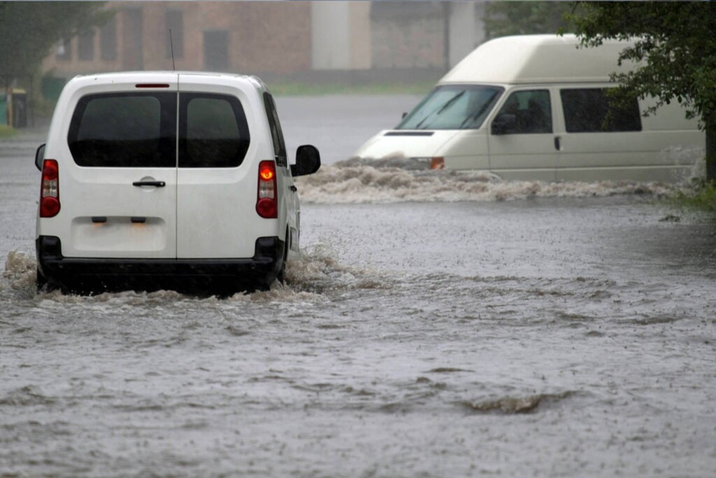 Dva bílé dodávkové vozy jedoucí po silnici zaplavené vodou.