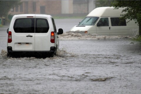 Dva bílé dodávkové vozy jedoucí po silnici zaplavené vodou.