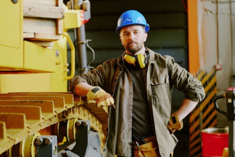 Pracovník ve stavební modré přilbě a pracovních rukavicích se opírá o žluté podvozkové části stavebního stroje v dílně.